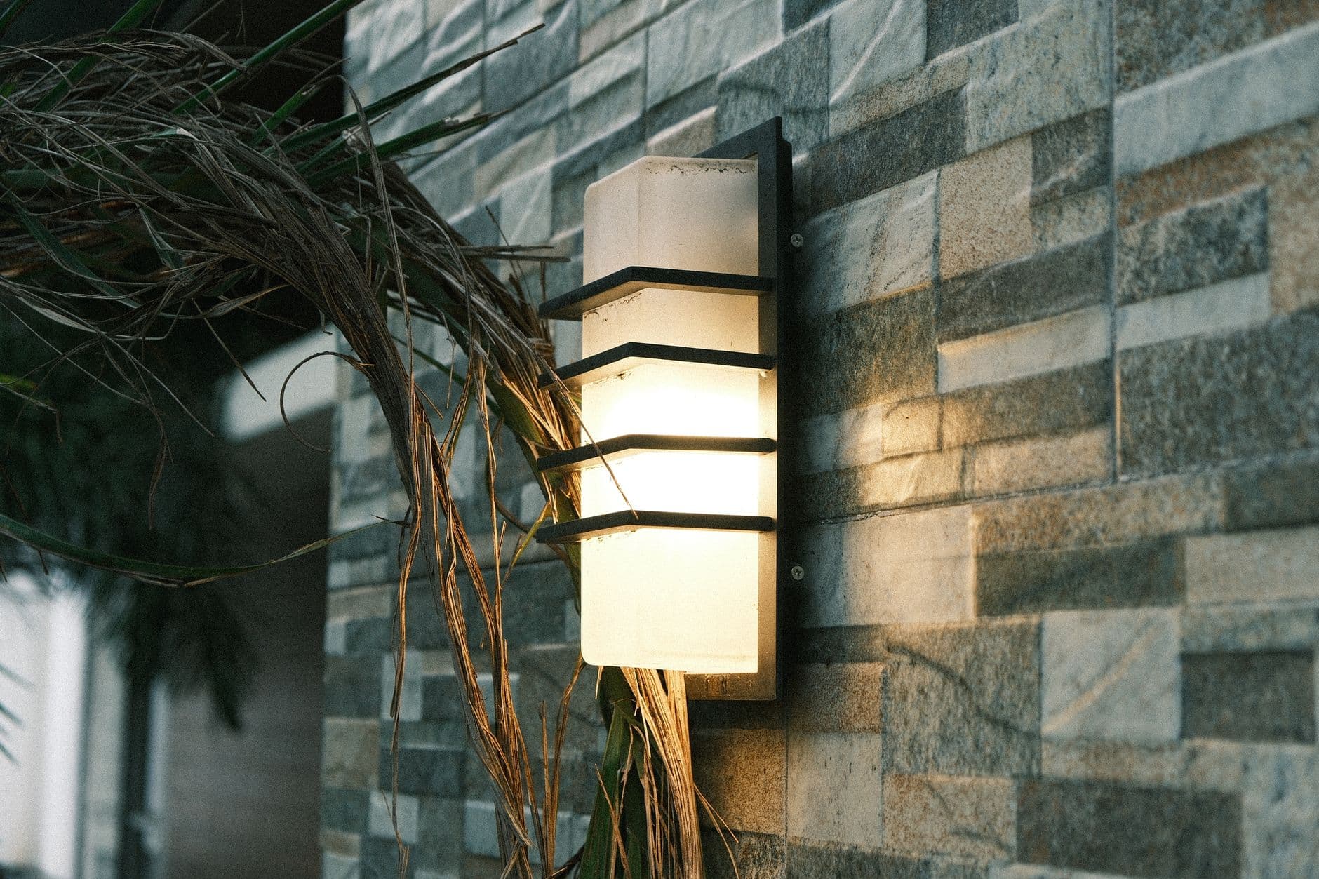 A lit outdoor wall light on a stone-clad wall beside planting.